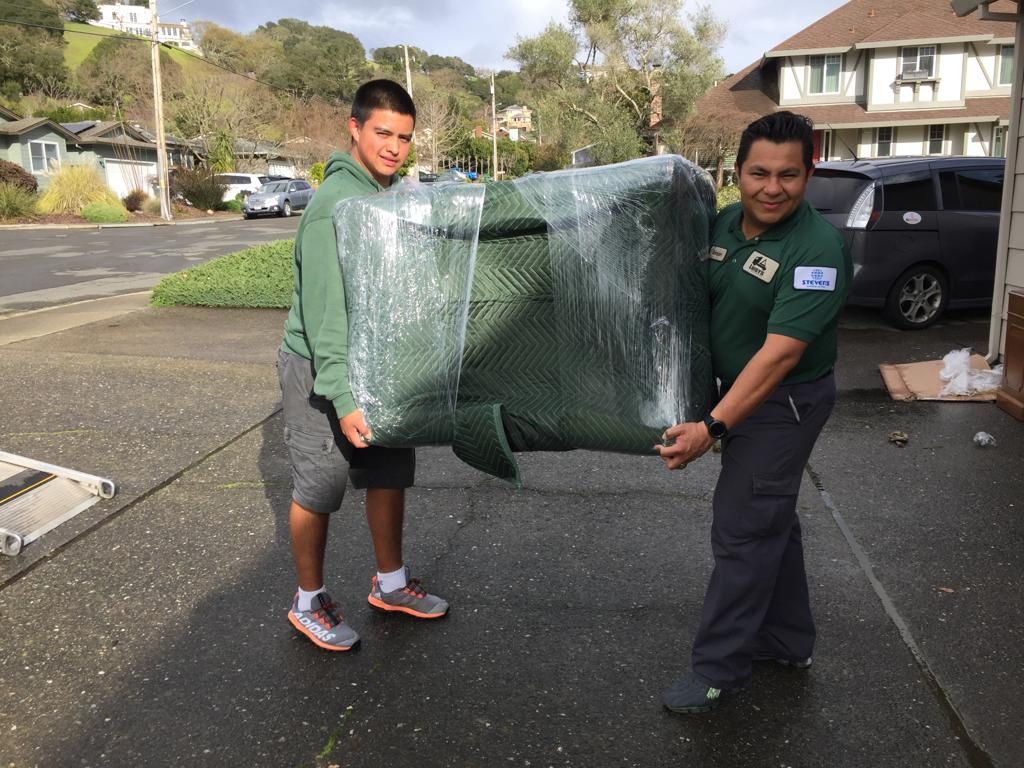 Two men wearing green shirts carrying a large green chair